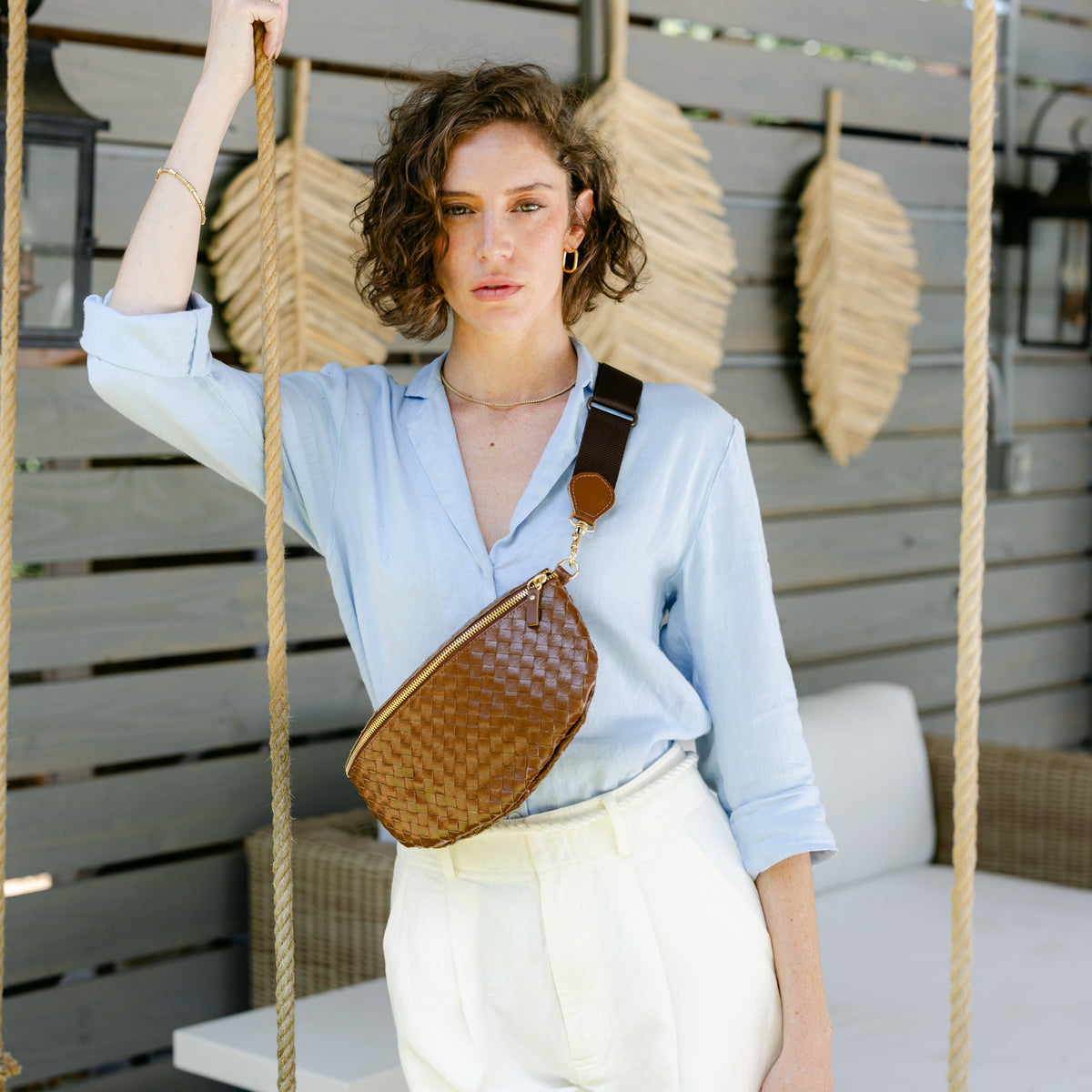 A woman wearing a light blue button-down shirt and white shorts stands on a porch swing holding a rope, wearing a brown woven crossbody bag with a brown leather strap.